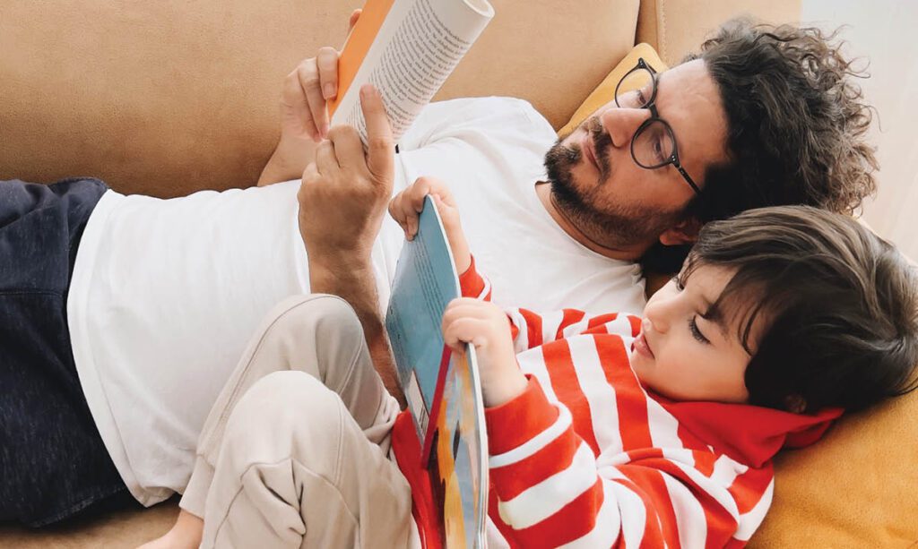 A man and a child reading a book on a couch.